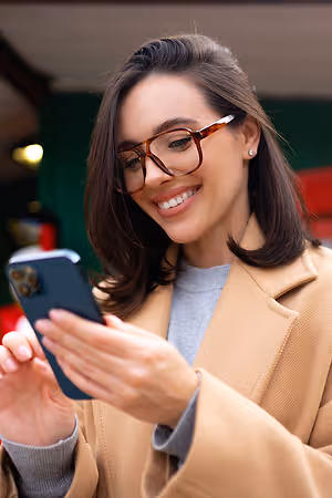 A woman wearing glasses and a beige coat is smiling while looking at her cell phone