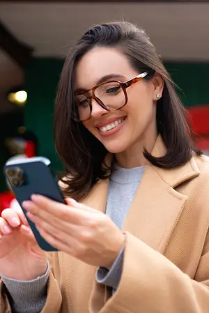 A woman wearing glasses and a beige coat is smiling while looking at her cell phone