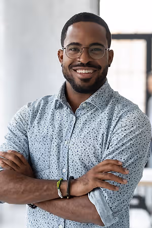 A smiling adult man with glasses and a bracelet on his left wrist, folded arms and standing in front of a white wall and a glass window. 