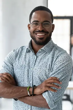 A smiling adult man with glasses and a bracelet on his left wrist, folded arms and standing in front of a white wall and a glass window. 