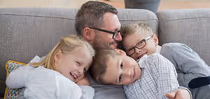 A man and his three children laying on a couch together smiling at the camera.