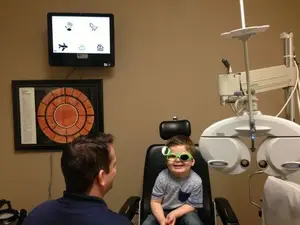 A man and a boy are sitting in a room with medical equipment, the boy is wearing sunglasses and the man is looking at him. Behind them, there is a monitor mounted on the wall with a picture frame, and a medical machine is placed in front of them.