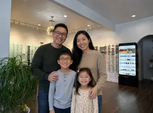 Family of three standing in front of an optometrists display of glasses with a large display case of glasses behind them