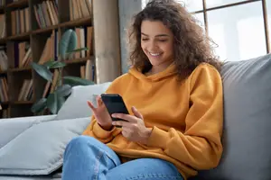 A smiling woman sitting on a couch, holding a cell phone and looking at it