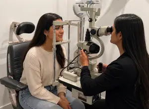 An adult woman is sitting in a chair and getting her eyes examined by another woman who is holding an eye exam tool.
