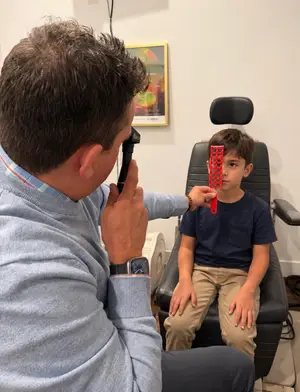 A man is examining a young boy's eyes using an ophthalmoscope in a clinic setting.