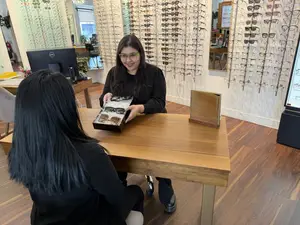 A woman holding a box of sunglasses is standing next to another woman sitting at a desk in a store.