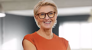 A woman with short blonde hair wearing glasses and a red shirt is smiling in front of a wall with a light coming through a window