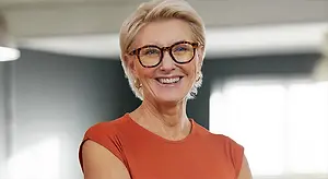 A woman with short blonde hair wearing glasses and a red shirt is smiling in front of a wall with a light coming through a window