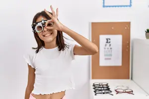 A young girl is standing in front of a white wall with her eyes closed, holding an eyeglass frame, and her hand raised in front of her face.