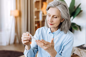An older woman is sitting on a couch in a living room and holding a bottle in her hand.