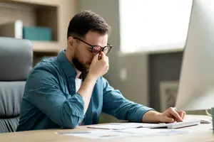A man is sitting at a desk and seems to be working on his computer, his eyes are closed and his hand is on his forehead.
