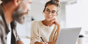 A man and a woman are sitting together, with the woman wearing glasses and looking at the man, who is smiling and looking at her. The woman is holding a pen and looking at a laptop, while the man is holding a cup. Behind them is a blurry background of a white wall and a window.