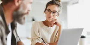 A man and a woman are sitting together, with the woman wearing glasses and looking at the man, who is smiling and looking at her. The woman is holding a pen and looking at a laptop, while the man is holding a cup. Behind them is a blurry background of a white wall and a window.