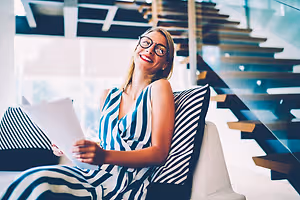A smiling woman wearing glasses is sitting on a couch and holding a piece of paper