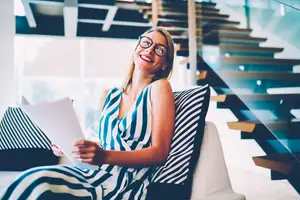 A smiling woman wearing glasses is sitting on a couch and holding a piece of paper