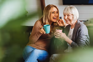 Two women sitting on a couch, one holding a cup and smiling, and the other woman smiling and looking at the cup