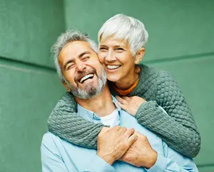 A smiling elderly man and woman standing close to each other and hugging each other in front of a green wall