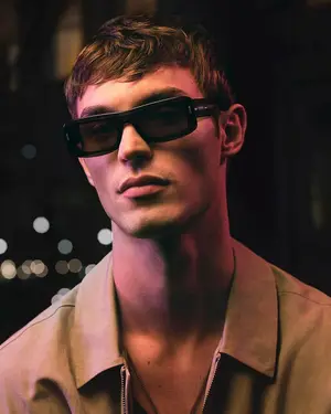 A young man wearing sunglasses and a tan shirt is standing and posing for a picture.