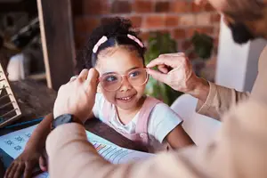 A girl wearing glasses is being helped by a man in front of a table with a book and a plant.