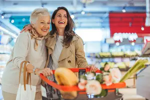Two women smiling and standing in a grocery store with a shopping cart