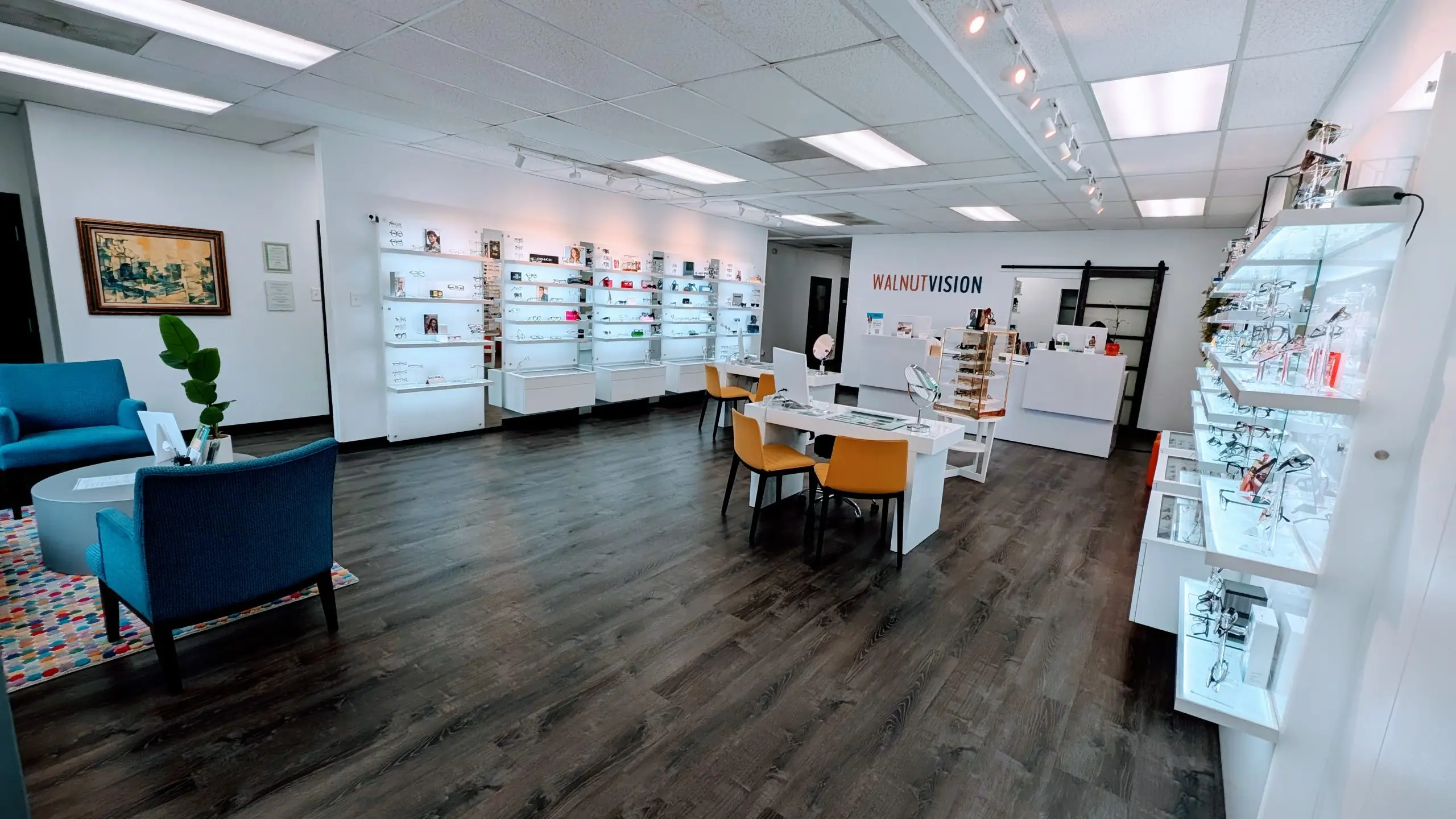 A clean and bright interior of a Walnut Vision optical store with a large white wall and ceiling lights and a white floor, white furniture, and a table with chairs and a potted plant.