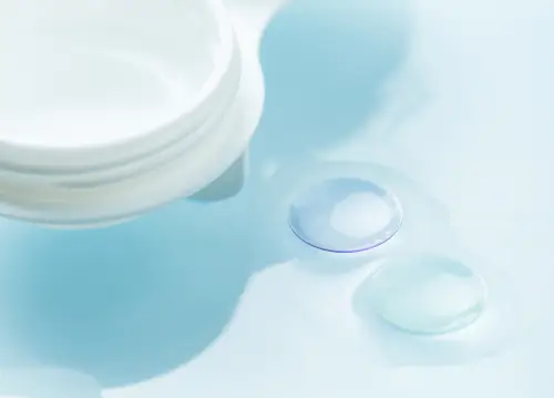 Close up of a container of contact lens solution with three lenses on a blue surface.