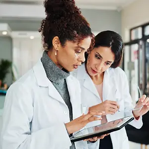 Two doctors in white lab coats are reviewing a tablet, with one woman holding a pair of glasses. They are in a medical office setting.