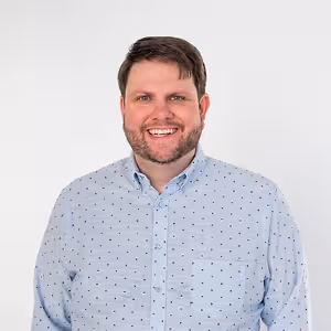 A smiling man wearing a light blue shirt with small multicolored dots against a white background