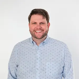 A smiling man wearing a light blue shirt with small multicolored dots against a white background