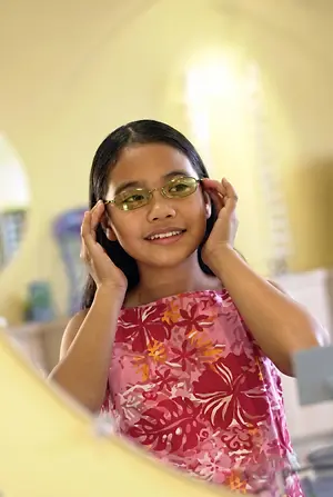 A girl with glasses and a floral dress stands in front of a mirror, smiling and adjusting her glasses.