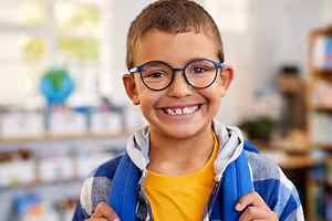 A boy wearing glasses and a blue hoodie smiles and poses for a photo in a classroom