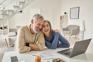 A couple sitting at a table with a laptop and papers, smiling and looking at the laptop.