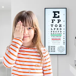 A young girl wearing an orange and white striped shirt stands in front of an eye exam chart, squinting with one hand over her eyes.