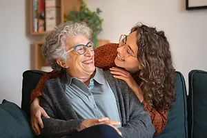 An elderly woman with glasses is sitting on a couch with a smiling young woman, both of them wearing glasses and looking happy