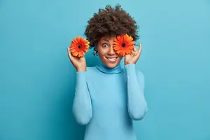 A woman is smiling and holding two orange flowers in her hands and posing for a photograph.