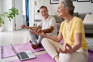A couple sitting on yoga mats smiling at each other with a laptop between them
