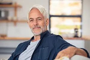 A mature man with a white beard and gray hair sitting on a couch and smiling at the camera