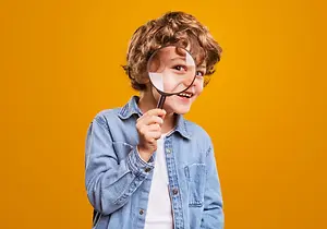 A smiling young boy with curly hair is holding a magnifying glass and looking through it while wearing a denim jacket and white shirt against a yellow background.