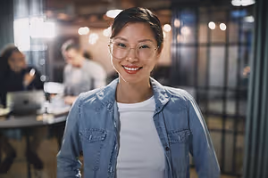 A woman in a denim jacket and glasses is smiling in an office setting.