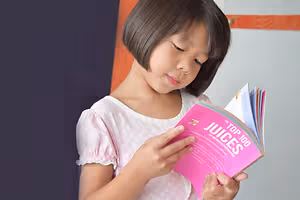 A young girl with short hair wearing a white polka-dot dress is holding a book titled 'The Top 100 Juices'.