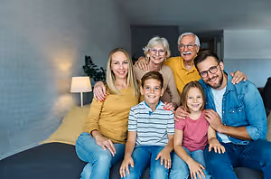 A family of six people, including two grandparents, a mother, a father, and two children, are sitting on a bed in a room with white brick walls, a lamp, and a plant, posing for a family photo.