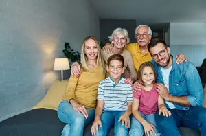 A family of six people, including two grandparents, a mother, a father, and two children, are sitting on a bed in a room with white brick walls, a lamp, and a plant, posing for a family photo.