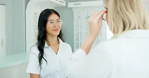 A young woman with glasses is smiling and looking in a mirror while a woman in a white shirt is adjusting her glasses in a hospital room.