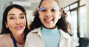 A smiling woman and a girl wearing glasses are standing together in a room with glass windows and a white wall.