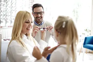 Three people wearing white lab coats smile in an optometrist's office, two women and a man with glasses, with the man holding a pair of glasses in his hands.