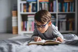 A young boy wearing glasses is lying on a bed and reading a book with his right hand.