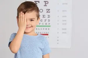 A smiling boy with brown hair wearing a blue t-shirt is looking at something with his right eye closed in front of an eye chart