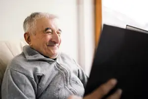 An elderly man is sitting on a chair and reading a book with a smiling face