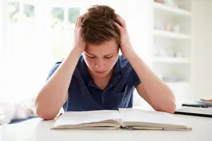 A boy sitting at a table with an open book is holding his head in his hands.